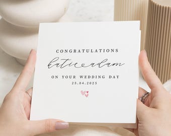 Op de afbeelding: Witte kaart met zwarte kalligrafie die "Gefeliciteerd Katie & Adam met jullie huwelijk 25.04.2025" luidt. Twee roze harten bevinden zich onder de tekst.