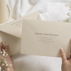 Op de afbeelding: Twee crèmekleurige enveloppen met zwarte bedrukte tekst. De voorste envelop toont de naam AMBER HENDERSON en een adres in Londonderry, Noord-Ierland. De geopende achterste envelop bevat de tekst KINDLY RESPOND en een retouradres.