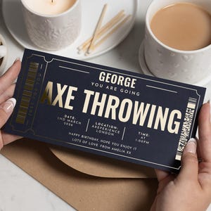 Op de afbeelding: Een marineblauw ticket met gouden letters voor "AXE THROWING" wordt vastgehouden. Het ticket bevat de naam van de ontvanger, datum, tijd en locatie. Een kaars, lucifers en een kopje thee staan op de achtergrond.