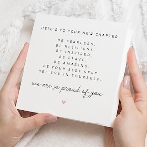 Op de afbeelding: Een vierkante, gebroken witte kaart, vastgehouden door handen, met zwarte tekst met de tekst "HERE'S TO YOUR NEW CHAPTER" gevolgd door inspirerende zinnen. Een klein roze hartje staat onderaan, met de woorden "we are so proud of you."