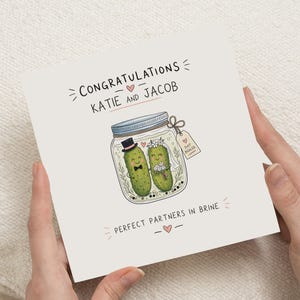 Peut inclure: Une carte de vœux carrée sur fond blanc avec une illustration de deux cornichons verts dans un bocal en verre. Le texte indique CONGRATULATIONS KATIE AND JACOB et PERFECT PARTNERS IN BRINE. Un cornichon porte un haut-de-forme et un nœud papillon, l'autre une couronne de fleurs et un voile.