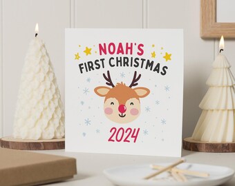 Op de afbeelding: Een witte kaart met rode en blauwe tekst die "Noah's First Christmas 2024" zegt. Een cartoonrendier met een rode neus bevindt zich in het midden van de kaart. De kaart is omringd door witte sneeuwvlokken en gele sterren.