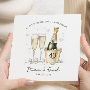 May include: A square card with a champagne illustration for a ruby wedding anniversary. The design includes two champagne flutes, a bottle in an ice bucket, and the text "Happy Ruby Wedding Anniversary". The card also has the text "Mum & Dad 1986 2026".
