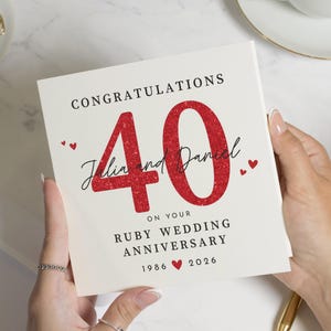 Op de afbeelding: Een witte kaart met de tekst "Gefeliciteerd 40" in zwart en rode glitter. De kaart luidt "Julia en Daniel op uw Robijnen Huwelijksverjaardag 1986 2026" met kleine rode hartaccenten. De kaart wordt door een persoon vastgehouden.