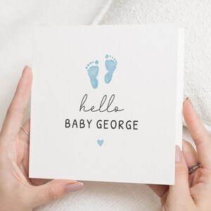 Könnte beinhalten: Eine weiße Karte mit blauen Baby-Fußabdrücken, dem Wort "hello" in Schreibschrift und dem Text "BABY GEORGE" in fetter Schrift. Unter dem Text befindet sich ein kleines blaues Herz. Die Karte wird von zwei Händen gehalten.