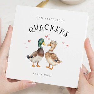 May include: A square greeting card with the text "I AM ABSOLUTELY QUACKERS ABOUT YOU!" features a watercolour illustration of two ducks. One duck has a green head and the other wears a floral crown. Small pink hearts are scattered around the ducks.
