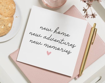 May include: A white card with the text "new home new adventures new memories" written in black cursive. A small pink heart is below the text. The card is on top of a pink notebook with a gold pen.