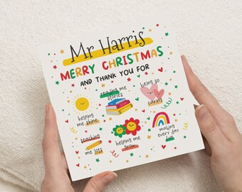 以下が含まれることがあります： 「Mr Harris MERRY CHRISTMAS AND THANK YOU FOR」のテキストと様々なイラストが描かれたクリスマスカード。太陽、本、ハート、花、虹が含まれています。カードは両手で持たれています。