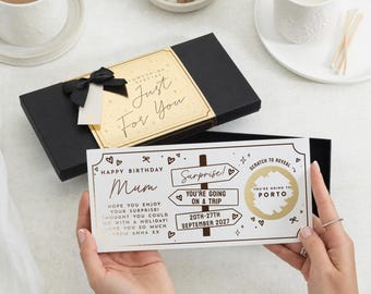 Op de afbeelding: Een zwarte geschenkdoos met een gouden Just For You label en een zwarte strik, vergezeld van een witte kras-verjaardagskaart voor mama. De kaart bevat een wegwijzer-afbeelding, de tekst You're Going On A Trip en een gouden cirkel die Porto onthult.