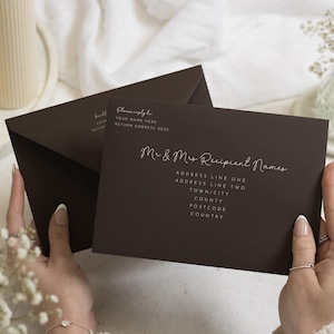 Op de afbeelding: Een donkerbruine rechthoekige envelop met witte bedrukte tekst. De voorzijde toont "Please reply to," "YOUR NAME HERE," "RETURN ADDRESS HERE," en "Mr & Mrs Recipient Names" gevolgd door tijdelijke aanduidingen voor adresregels, stad, postcode en land.
