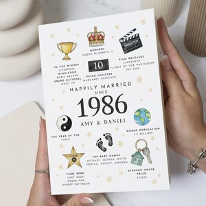 Op de afbeelding: Een witte wenskaart met de titel Happily Married Since 1986 voor Amy en Daniel. Het bevat illustraties van een kroon, trofee, filmklapper, yin-yang, wereldbol, babyvoetjes, ster en huissleutel, met tekst over historische feiten uit 1986.