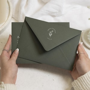 Op de afbeelding: Een donker olijfgroene envelop met een witte florale lijntekening, de namen Amelia & Nathan en de datum 08.05.27 gedrukt op de flap. De envelop wordt dichtgehouden en bedekt gedeeltelijk een bijpassende kaart met zichtbare tekst.