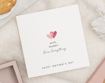 May include: A white Mother's Day card with the words "Wife, Mummy, Our Everything" and "Happy Mother's Day" in black script. Two watercolor hearts in pink and red are above the text. The card is on a white surface.