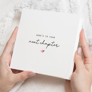 May include: A square, cream-coloured card held by hands, with the text "Here's to your next chapter" in a handwritten font. Two small pink hearts are below the text. The card is on a white, textured surface.