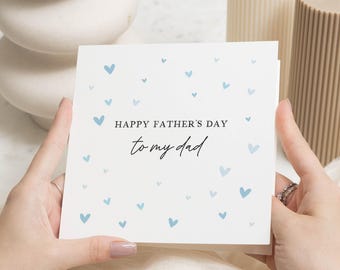 May include: A white Father's Day card with blue heart accents and the message "Happy Father's Day to my dad" in black script. The card is held by hands, with a ring visible on one finger.