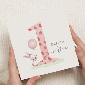 May include: A square birthday card with a large number "1" adorned with strawberries. A charming bunny holds a pink balloon. The text "OLIVIA is One" is written in black script.