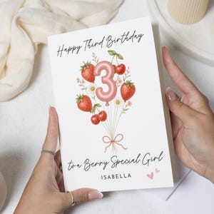 May include: A white greeting card featuring the text Happy Third Birthday at the top and to a Berry Special Girl at the bottom. The center displays a pink balloon shaped like the number 3, surrounded by watercolor strawberries, cherries, and flowers.