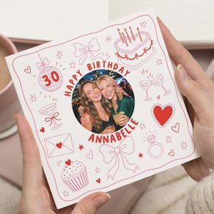 Op de afbeelding: Een vierkante verjaardagskaart met een centrale ronde foto van twee personen. De kaart bevat rode lijntekeningen van een taart, strikken, een discobal met het getal 30, hartjes, een cupcake en een ring. De tekst luidt Happy Birthday Annabelle.
