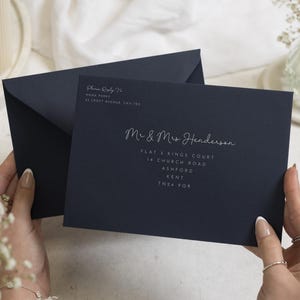 Op de afbeelding: Een donkerblauwe envelop met witte bedrukte tekst die luidt Please Reply To Anna Parks 41 Croft Avenue, CA1 1BG, en Mr & Mrs Henderson, Flat 3 Kings Court, 14 Church Road, Ashford, Kent, TN24 9QR, met een bijpassende open envelop erachter.