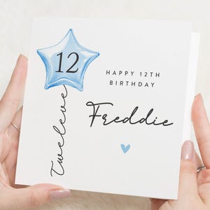 Könnte beinhalten: Eine weiße Geburtstagskarte mit einem blauen sternförmigen Ballon mit der Zahl 12. Die Karte trägt die Aufschrift "Happy 12th Birthday Freddie" in schwarzer Schrift. Ein kleines blaues Herz ist ebenfalls vorhanden.