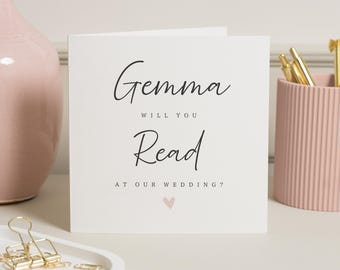 May include: A white card with the text "Gemma will you Read at our wedding?" in black script. A small pink heart is at the bottom. The card is on a white surface with a pink vase and pen holder in the background.
