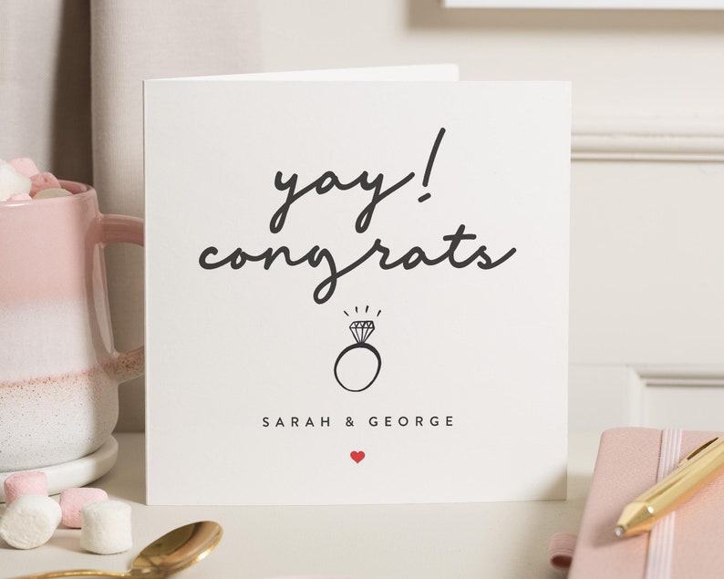 Może przedstawiać: Kwadratowa biała kartka z czarnym napisem "yay! congrats". Pod tekstem znajduje się ilustracja pierścionka z diamentem. Pod pierścionkiem wydrukowane są imiona "SARAH & GEORGE", a pod nimi małe czerwone serce.