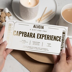 Könnte beinhalten: Ein cremefarbenes Ticket mit goldfarbenen Buchstaben, auf dem "CAPYBARA EXPERIENCE" und "ALICIA, A GIFT FOR YOU" steht. Das Ticket enthält ein Datum, einen Ort und eine Uhrzeit. Das Ticket wird vor einer Marmoroberfläche mit einer Tasse Tee, einer Kerze und einer Schale mit Zuckerwürfeln gehalten.