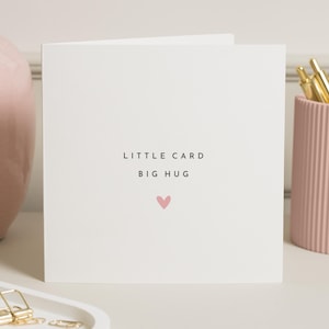 Può includere: Un biglietto di auguri bianco con la scritta "LITTLE CARD BIG HUG" e un piccolo cuore rosa. Il biglietto è appoggiato su una superficie bianca, con un vaso rosa e un portapenne sullo sfondo.