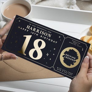 Könnte beinhalten: Eine marineblaue Geburtstagskarte mit goldenen Folienakzenten, gehalten von einer Person. Die Karte lautet "Happy 18th Birthday Harrison" mit einer großen "18" und einem Rubbelfeld für Fahrstunden.