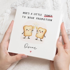 Peut inclure: Carte de vœux blanche avec le texte "Here's a little toast to your promotion" et le nom "Oscar". La carte présente des illustrations de deux tranches de pain grillé tenant des verres de champagne. Un petit cœur rouge est en dessous du nom.