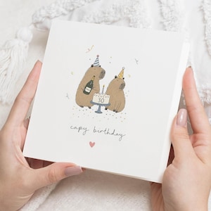 Op de afbeelding: Een vierkante witte wenskaart met een cartoonillustratie van twee bruine capibara's met feesthoedjes. De een houdt een champagnefles vast, de ander zit bij een taart met het getal 30. De tekst luidt capy birthday met een klein roze hartje.