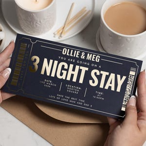 May include: A navy blue ticket with gold lettering and accents, held by two hands. The ticket reads "Ollie & Meg, 3 Night Stay" with date, location, and time details. A cup of tea, a candle, and sugar cubes are in the background.