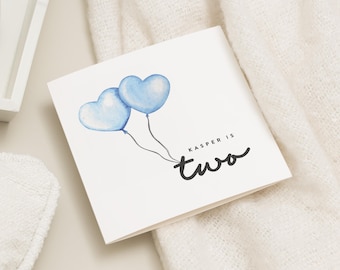 May include: A white card with two blue heart-shaped balloons and the text "Kasper is two" in black cursive writing.
