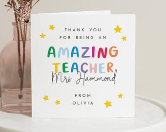 Puede incluir: Una tarjeta blanca con el texto "THANK YOU FOR BEING AN AMAZING TEACHER Mrs Hammond FROM OLIVIA" en letras coloridas. Ilustraciones de estrellas amarillas rodean el texto. La tarjeta está sobre una superficie blanca.