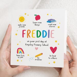 Może przedstawiać: Kwadratowa kartka z imieniem "FREDDIE" w kolorowych literach, otoczona ilustracjami słońca, jabłka, książek, tęczy, serca i ołówka. Tekst brzmi "on your first day at Kingsley Primary School."