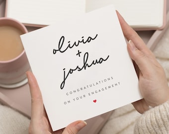 May include: White card with black calligraphy that reads "Olivia + Joshua" and "Congratulations on your engagement" with a small red heart.