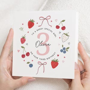 May include: A square white greeting card featuring a large pink number 3 and the name Olivia in black script. The design includes illustrations of strawberries, cherries, raspberries, blueberries, and pink bows, with the text To a berry special girl on your third birthday.