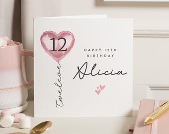 Op de afbeelding: Een witte verjaardagskaart met een roze hartvormige ballon met het nummer 12. De kaart leest "Happy 12th Birthday Alicia" in zwart schrift. Twee kleine roze hartjes onderaan.