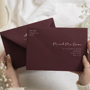 Könnte beinhalten: Zwei dunkelburgunderrote Umschläge mit weißem Textaufdruck. Der vordere Umschlag ist an Mr and Mrs Brown, 54 Victoria Road, Dundee, Scotland, DD1 2LA adressiert. Der hintere Umschlag zeigt eine Absenderadresse für Amelia Joyes in Leicester.