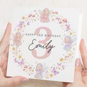 Può includere: Un biglietto di compleanno bianco con il numero 3 rosa e il nome "Emily" in corsivo. Il biglietto presenta illustrazioni di fate, farfalle, funghi e stelle, con il testo "Happy 3rd Birthday".