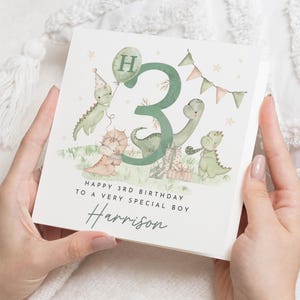 Könnte beinhalten: Quadratische Geburtstagskarte mit einer Dinosaurier-Illustration und der Zahl 3. Die Karte trägt die Aufschrift "Happy 3rd Birthday to a Very Special Boy" und enthält den Namen "Harrison". Die Karte wird von einer Person gehalten.