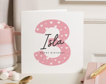 Peut inclure: Carte d'anniversaire blanche avec le prénom "Isla" en écriture noire et le chiffre "3" rose orné de cœurs blancs. Les mots "HAPPY BIRTHDAY" sont en dessous.