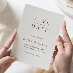 Pode incluir: Um cartão creme "Save the Date" com texto preto. O cartão diz "SAVE the DATE" acima dos nomes "EMMA & REECE" e da data "27TH SEPTEMBER 2027". O cartão é segurado por duas mãos.