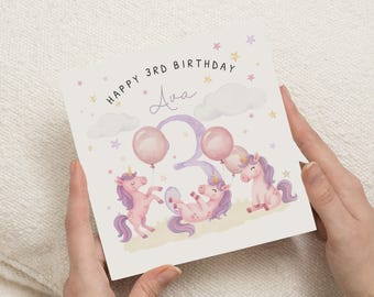 May include: A birthday card with the text "Happy 3rd Birthday Ava" and illustrations of unicorns, balloons, clouds, and stars. The unicorns are pink and purple, and the balloons are pink. The card is held by two hands.