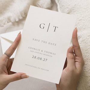 Op de afbeelding: Een crèmekleurige trouwuitnodigingskaart, vastgehouden door handen, met de initialen "G|T" bovenaan. De tekst "SAVE THE DATE" en de namen "GEORGIE & THOMAS" staan eronder gedrukt, samen met de datum "28.08.27". De woorden "INVITATION TO FOLLOW" staan onderaan.