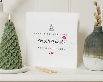 May include: A white greeting card with the words "Happy First Christmas married" and the names "Mr & Mrs Johnson". A green Christmas tree-shaped candle is lit on the left. A small vase with a plant is on the right.