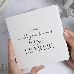 Peut inclure: Une carte blanche avec le texte "ZACHARY, will you be our RING BEARER?" en écriture noire. La carte est tenue dans les mains avec une bague au doigt. Une enveloppe blanche est visible derrière la carte.