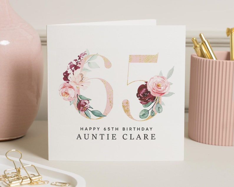 May include: A white card with a gold and pink floral design featuring the number 65. The text "Happy 65th Birthday Auntie Clare" is printed below the floral design.