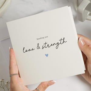 May include: A square greeting card with the words "Sending you love & strength" in black script, with a small blue heart. The card is held by a person. A white mug and a white bowl are in the background.