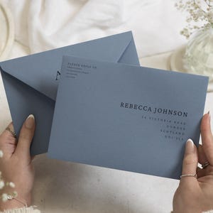 Op de afbeelding: Een stoffig blauwe envelop met de gedrukte naam REBECCA JOHNSON, een straatadres en een retouradresgedeelte met het label PLEASE REPLY TO. De envelop heeft een gladde matte afwerking en donkerblauwe schreefloze typografie in het midden.